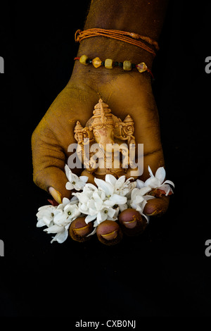 Hinduistische Elefantengott. Kurkuma bedeckt indischen mans Hand mit Lord Ganesha Statue und Jasmin Blüten auf schwarzem Hintergrund Stockfoto