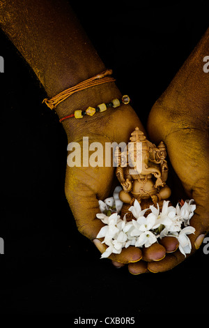 Hinduistische Elefantengott. Kurkuma bedeckt indischen mans Hand mit Lord Ganesha Statue und Jasmin Blüten auf schwarzem Hintergrund Stockfoto