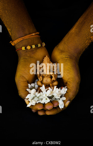 Hinduistische Elefantengott. Kurkuma bedeckt indischen mans Hand mit Lord Ganesha Statue und Jasmin Blüten auf schwarzem Hintergrund Stockfoto