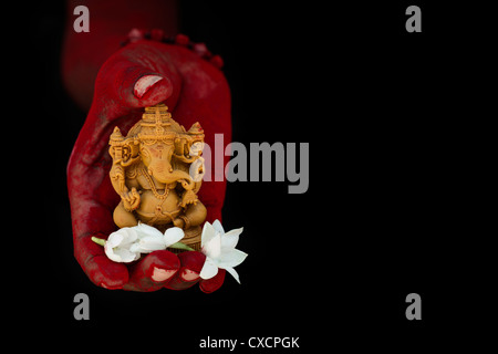 Hinduistische Elefantengott. Rotes Pulver bedeckt indischen mans Hand mit Lord Ganesha Statue und Jasmin Blüten auf schwarzem Hintergrund Stockfoto