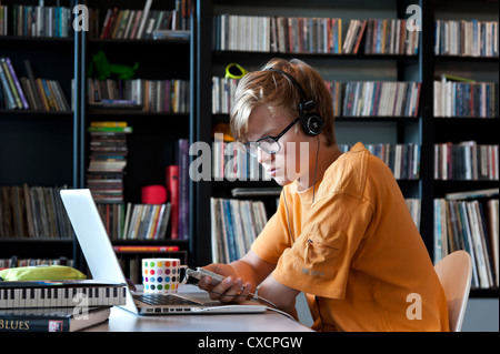 Studious entspannt teenage Schule Jungen 14-16 Jahre Kopfhörer trägt mit seinem Apple iPhone Smartphone und Laptop in der Musik Studium Bibliothek Stockfoto