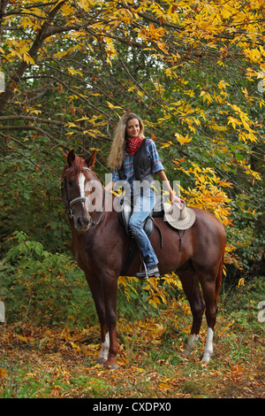 Cowgirl trägt ein kariertes Hemd und Weste Reiten Reiten durch einen Wald mit herbstlichen Bäume Stockfoto