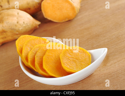 Gekochte Süßkartoffel (Ipomoea Batatas lat.) schneiden Sie in Scheiben in weiße Schüssel auf Holzoberfläche mit rohen Süßkartoffeln in den Rücken Stockfoto