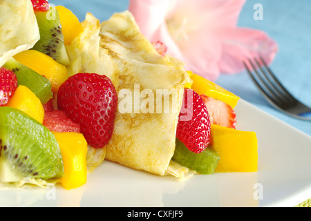 Crêpes gefüllt mit frischen Früchten (Erdbeeren, Kiwi, Mango, Wassermelone) mit einem rosa Gladiolen und einer Gabel im Hintergrund Stockfoto