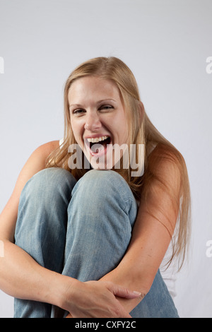 Schöne blonde Frau sitzt auf einem hölzernen Stuhl mit ihrem Knie in der Nähe von ihr Kinn, hob mit einem Lachen in die Kamera schaut Stockfoto