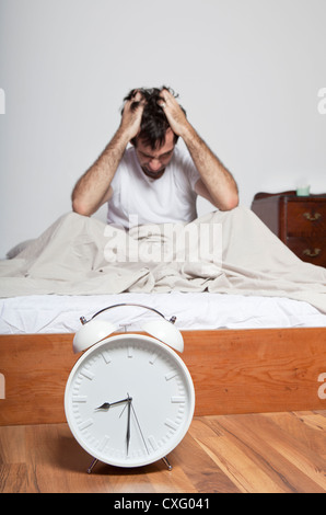 Ein junger müde Mann sitzt auf einem Bett und hielt seinen Kopf. Großer Wecker ist vor dem Bett platziert. Stockfoto