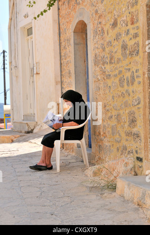 Ältere Dame, die vor ihrem Haus in einer schattigen Gasse sitzt und eine Zeitung liest, im Dorf Pano Elounda auf Kreta, griechische Insel, Griechenland Stockfoto