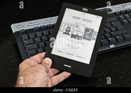 Ein Mann hält und liest eine Kobo Touch Ereader (Ebook) in der Nähe einer Computertastatur. Stockfoto