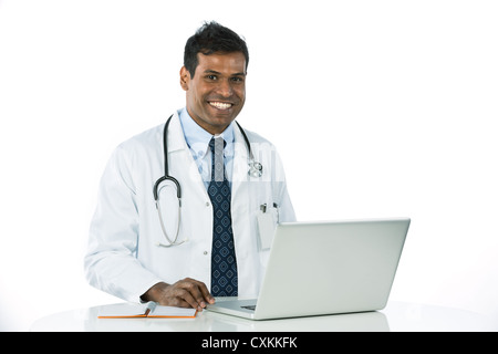 Männlicher indischer Arzt trägt einen weißen Mantel, sitzen an einem Schreibtisch arbeiten mit seinem Laptop. Stockfoto