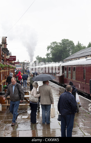 Passagiere auf der Plattform der Bury Bolton Street Station an einem regnerischen Tag wo eine Heywood-gebundenen Dampf Erbe trainieren ist Stand. Stockfoto