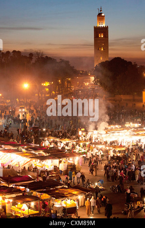 Marktstände und Menschen in der Abenddämmerung in Fna Jamaa el Fna Platz mit Souk Koutoubia-Minarett-Moschee Stockfoto