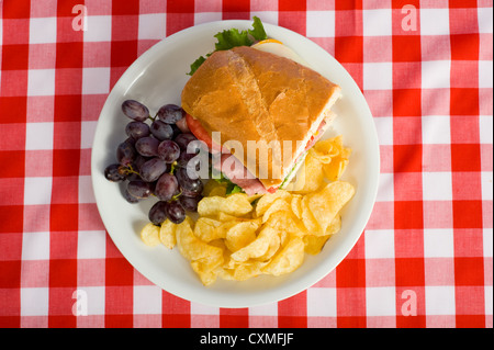 Ein Picknick-Mittagessen, bestehend aus einem Sandwich, Kartoffelchips und Trauben auf eine rote karierte Tischdecke Stockfoto