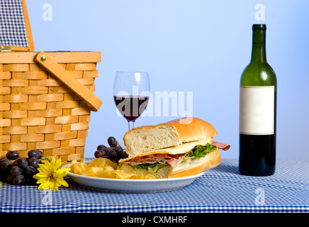 Ein Picknick-Mittagessen, bestehend aus einem Sandwich, Kartoffelchips und Trauben auf einer blau karierte Tischdecke mit einer Flasche Wein Stockfoto