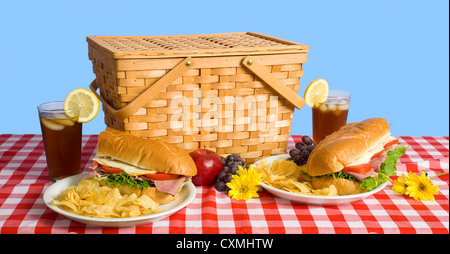 Ein Picknick-Mittagessen, bestehend aus einem Sandwich, Kartoffelchips und Trauben auf eine rote karierte Tischdecke mit Eistee und eine Zitrone Stockfoto