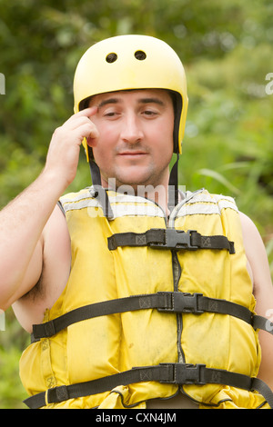 Ein Mann, der einen hellgelben Schutzhelm und eine passende gelbe Schwimmweste trägt, wird von der Brust nach oben gezeigt. Er justiert den Helm mit der rechten Hand und berührt die Seite neben seinem Tempel. Das Leben Stockfoto