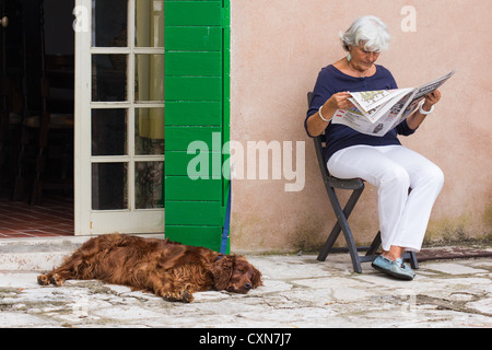 Frau, die draußen auf einem Stuhl sitzt und Zeitung hält und liest, mit einem roten Setter, der neben ihr liegt. Korcula. Stockfoto