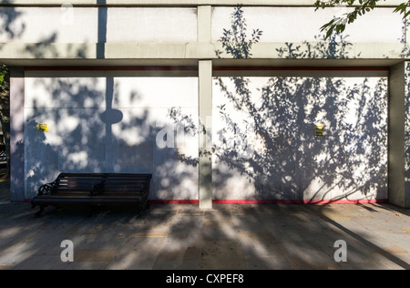 Eine Holzbank auf Pflaster gegen eine graue Wand mit scharfen Schatten der Bäume quer dazu stehen Stockfoto