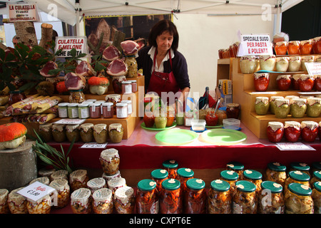 Eine Verkäuferin bietet typische Lebensmittel wie Schinken, Salami, Pestos, Saucen und Gurken auf dem Markt. Friaul, in Norditalien. Stockfoto