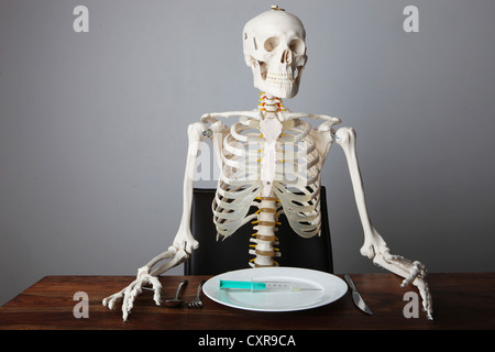 Skelett sitzen am gedeckten Tisch mit einer Spritze auf einem Teller Stockfoto