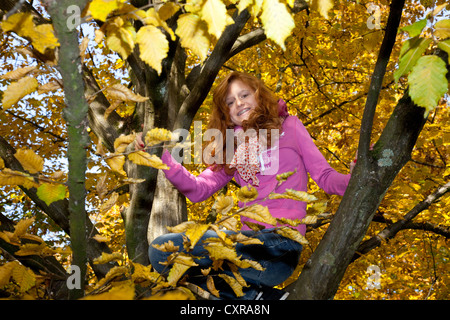 Rothaariges Mädchen mit Zahnspange sitzt in einem Baum mit herbstlichen Laub Stockfoto