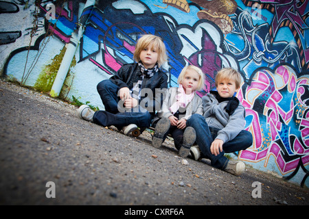 Geschwister, 2, 5 und 7 Jahren, sitzen auf einem Skateboard vor einer Wand mit graffiti Stockfoto