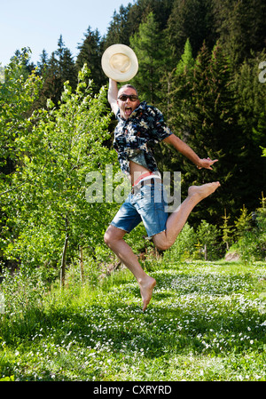 Mann mit Sonnenbrille und einem Hawaii-Hemd mit einem Strohhut auf einer blühenden Wiese springen lachen Stockfoto