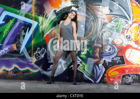 Junge Frau in einem kurzen grauen Kleid, schwarze Strümpfe und high Heels posiert vor einer Graffitiwand Stockfoto