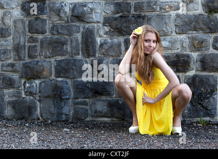 Junge Frau in einem gelben Kleid und eine gelbe Blume posiert in eine hockende Position vor einer Steinmauer Stockfoto