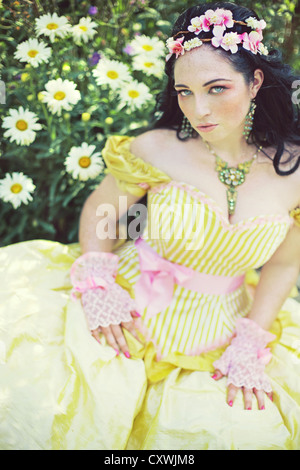 Eine junge Brünette Frau mit Blumen im Haar tragen fancy gelbes Kleid im Sommer sitzen von Gänseblümchen Stockfoto