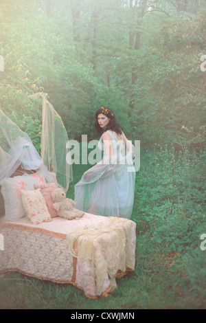 Ein hübsches Mädchen mit dunklen Haaren in ein schickes blass blauen Nachthemd abwendet ein kleines Kind Bett sitzen im Wald Stockfoto