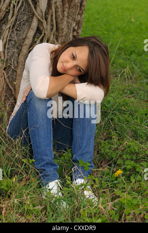 Nachdenkliche junge Frau der indonesischen Ursprungs, sitzen auf einer Wiese vor einem Baum Stamm, publicground Stockfoto