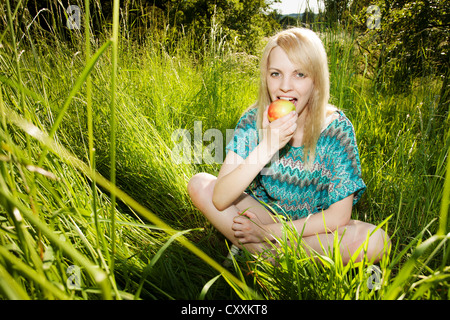 Lächelnde junge Frau sitzt in hohe Gräser Essen eines Apfels Stockfoto