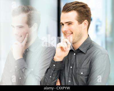 Lächelnde junge Geschäftsmann steht man vor einer Glasscheibe mit Reflexion Stockfoto