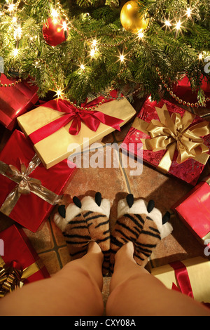 Person mit Hausschuhen in Form von Tiger Pfoten vor Weihnachtsgeschenke unter einem Weihnachtsbaum steht Stockfoto