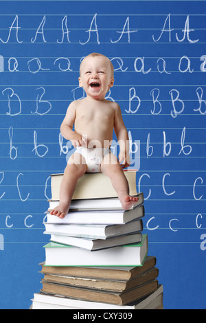 Lachendes Baby sitzt auf einem Stapel von Büchern vor einer Tafel mit Buchstaben darauf geschrieben Stockfoto