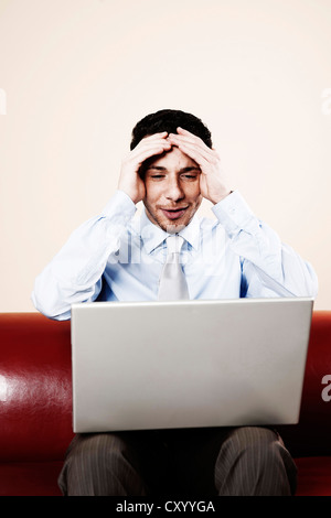 Junger Mann sitzt mit seinem Laptop auf dem Sofa, überrascht Stockfoto
