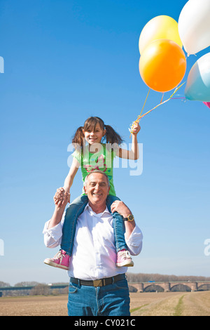 Mädchen mit vielen bunten Ballons in der Hand beim Sitzen auf den Schultern ihres Großvaters Stockfoto