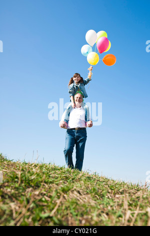 Mädchen mit vielen bunten Ballons in der Hand beim Sitzen auf den Schultern ihres Großvaters Stockfoto