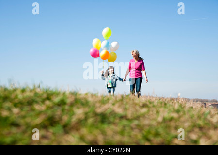 Mädchen mit Luftballons in der Hand während des Gehens mit ihrer Mutter über eine Wiese Stockfoto