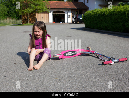 Kleines Mädchen, hat drei Jahre aus ihrem Roller, sitzen auf dem Boden Weinen verletzten Knie festhalten gefallen. Stockfoto