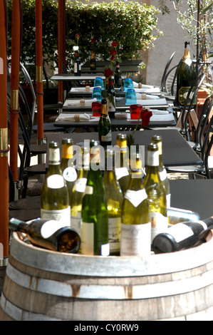 Leere Weinflaschen sitzen auf einem Fass vor einem Restaurant mit gedeckten Tischen im Hintergrund Stockfoto