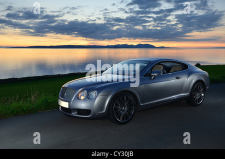 Bentley Continental GT neben Strand und Meer mit Sonnenuntergang und Inseln im Hintergrund und Wolken im Himmel Stockfoto