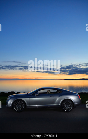 Bentley Continental GT neben Strand und Meer mit Sonnenuntergang und Inseln im Hintergrund und Wolken im Himmel Stockfoto