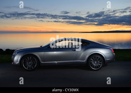 Bentley Continental GT neben Strand und Meer mit Sonnenuntergang und Inseln im Hintergrund und Wolken im Himmel Stockfoto