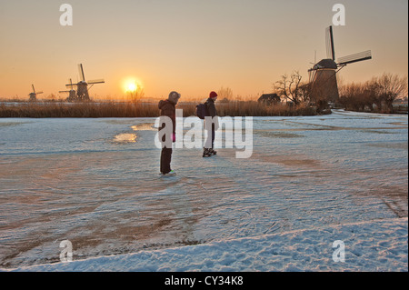 Eislaufen am zugefrorenen Kanal bei Sonnenuntergang Menschen passieren die Windmühlen von Kinderdijk in der Nähe von Rotterdam in den Niederlanden. Stockfoto