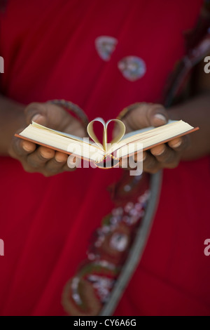 Indischer womans Hände halten eine Herzform aus Buch Seiten gegen roten Sari Hintergrund. Indien Stockfoto