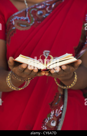 Indischer womans Hände halten eine Herzform aus Buch Seiten gegen roten Sari Hintergrund. Indien Stockfoto