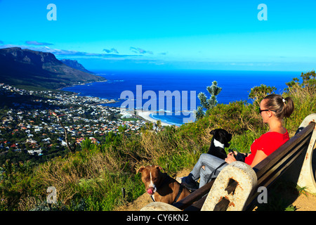 Ein junger weiblicher Tourist sitzt auf einer Bank mit Blick auf Camps Bay in der Nähe von Kapstadt in Südafrika. Stockfoto