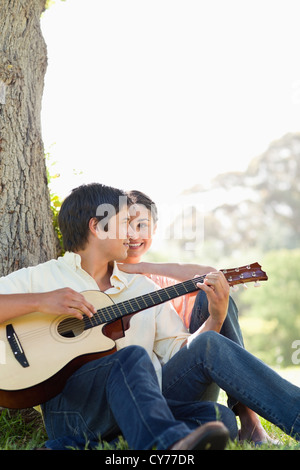 Mann, eine Gitarre zu spielen und gegen einen Baum zu sitzen, während sein Freund ihn Uhren Stockfoto