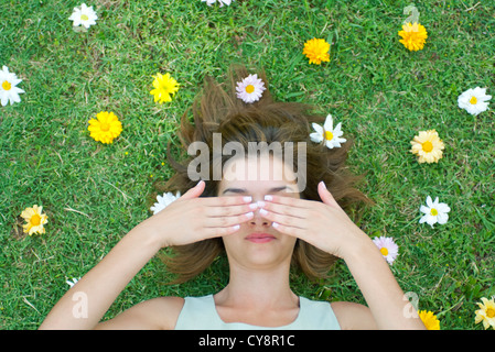 Frau liegt auf dem Rasen umgeben von Blumen mit Händen, Augen bedecken Stockfoto